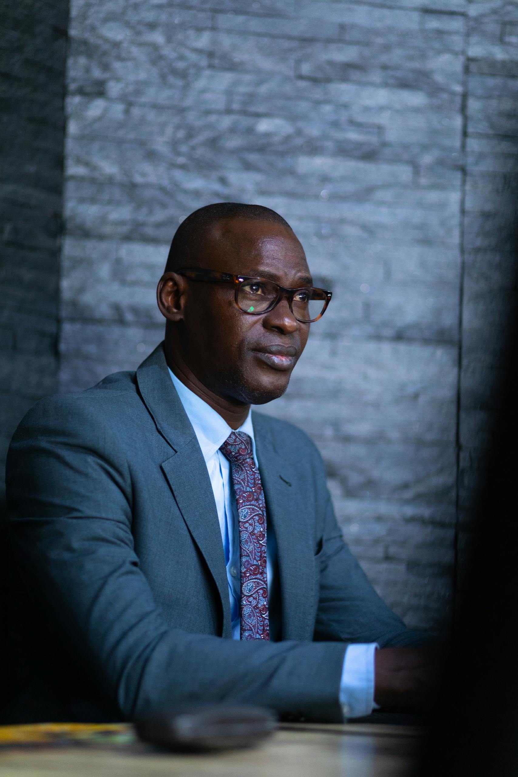 African American man in suit and glasses sitting against a stone wall, looking serious and professional.